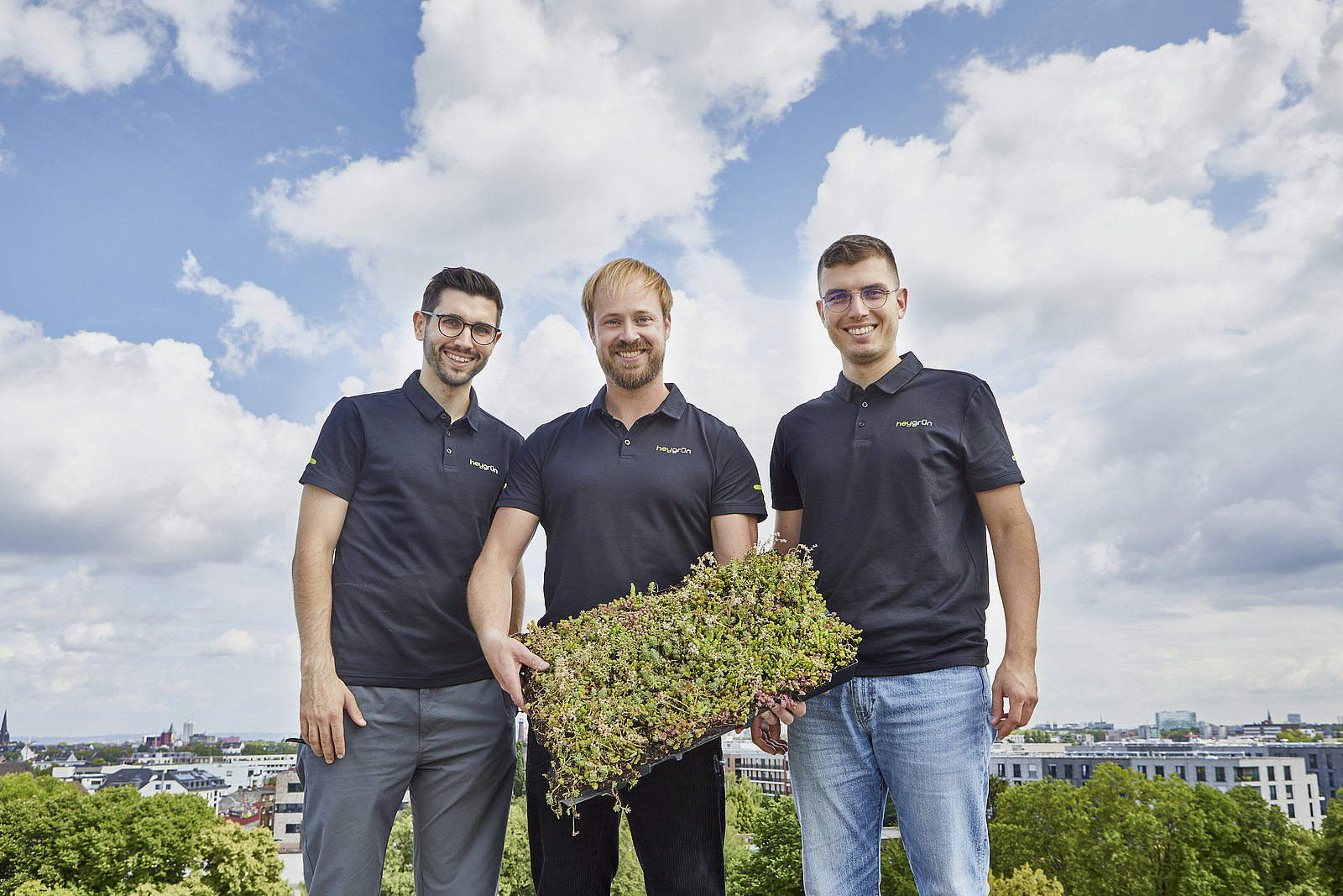 Teamfoto von heygrün. Drei junge Männer stehen lächelnd auf einem Dach. Einer hält ein Stück Dachbegrünung in der Hand.