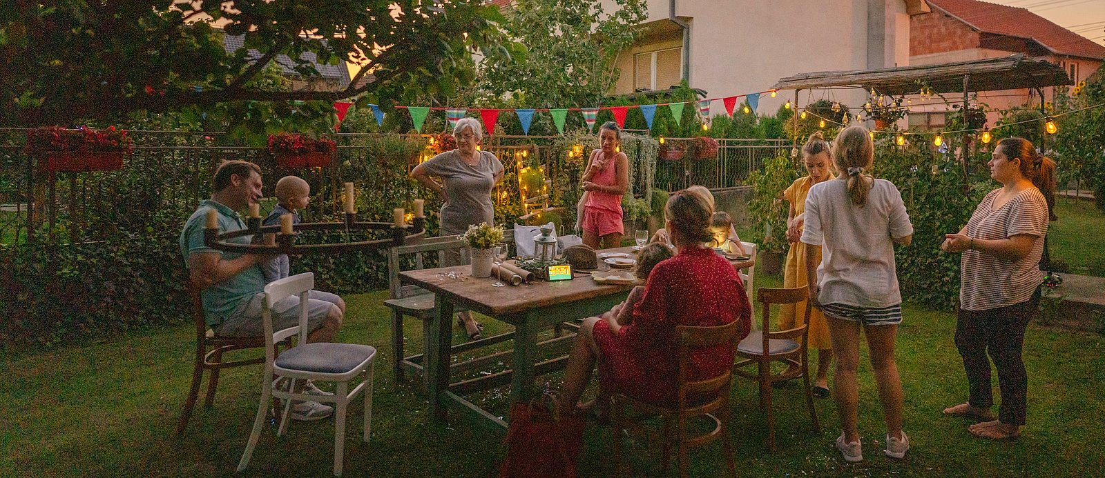 Menschen versammeln sich um einen Holztisch bei einer abendlichen Gartenparty.