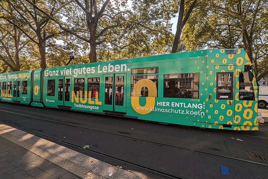 Foto der Kölner Klimabahn. Man sieht eine Straßenbahn der KVB, die mit vielen gelben Nullen bedruckt ist. Zudem trägt sie die Schriftzüge "Auf die Plätze, fertig Null" und "Ganz viel gutes Leben"