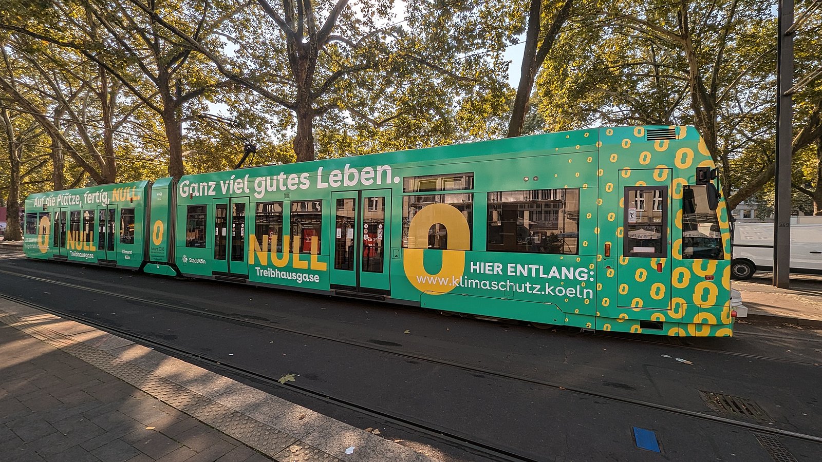 Foto der Kölner Klimabahn. Man sieht eine Straßenbahn der KVB, die mit vielen gelben Nullen bedruckt ist. Zudem trägt sie die Schriftzüge "Auf die Plätze, fertig Null" und "Ganz viel gutes Leben"